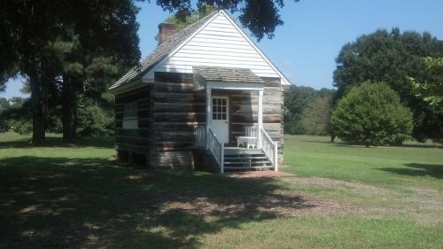 A small, rustic log cabin with a white wooden facade, surrounded by green grass and trees under a clear blue sky.