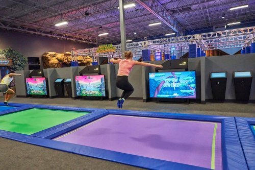 A person jumps on a trampoline in a vibrant indoor trampoline park with colorful mats and screens in the background.
