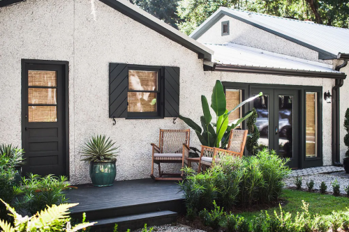 A cozy house exterior with dark shutters, a porch, and tropical plants, surrounded by greenery and a gravel path.