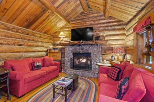 Cozy log cabin interior with red sofas, a stone fireplace, and a TV, featuring wooden beams and warm lighting.