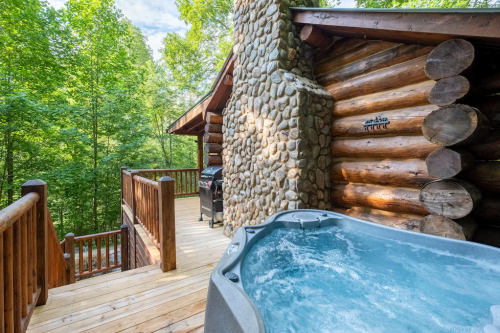 A wooden cabin with a stone exterior, featuring a hot tub and a deck surrounded by lush green trees.