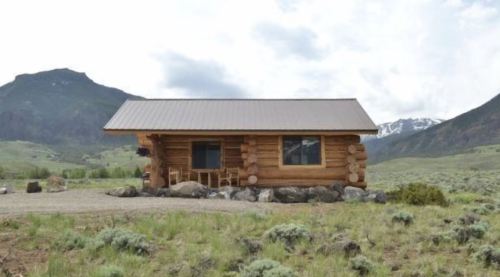 A rustic log cabin surrounded by mountains and greenery under a cloudy sky.