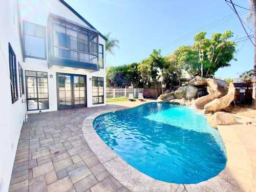 A sunny backyard with a pool, slide, and a two-story house surrounded by greenery and a stone wall.