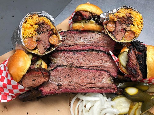 A platter featuring sliced brisket, two sandwiches, and wraps filled with macaroni and sausage, garnished with pickles and onions.