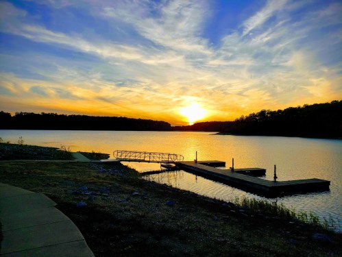 Sunset over a calm lake, with a dock and silhouettes of trees along the shore under a colorful sky.