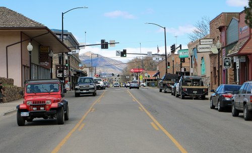 A street view featuring shops on either side, with vehicles driving and mountains in the background under a blue sky.