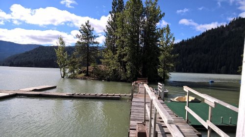 A wooden dock extends over a calm lake, surrounded by trees and mountains under a partly cloudy sky.