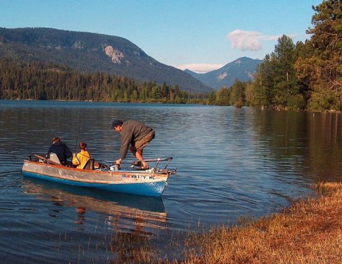 A man and two people in a small boat on a calm lake, surrounded by mountains and trees under a clear blue sky.