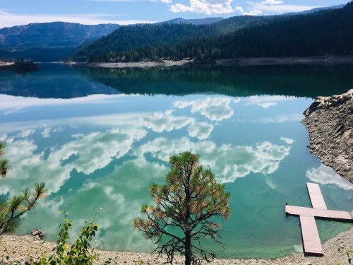 A serene lake reflecting clouds and trees, with a wooden dock extending into the water. Mountains in the background.