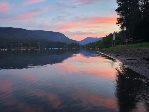 A serene lake at sunset, reflecting colorful skies and surrounding mountains, with trees lining the shore.
