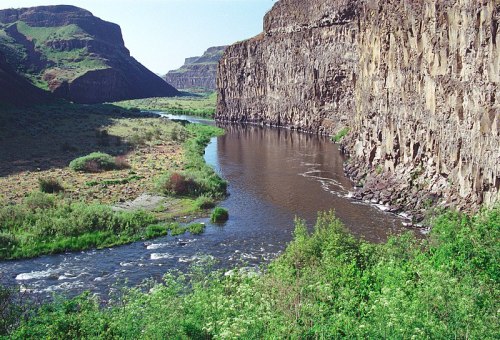 A winding river flows between steep, rocky cliffs, surrounded by lush greenery and rolling hills under a clear blue sky.