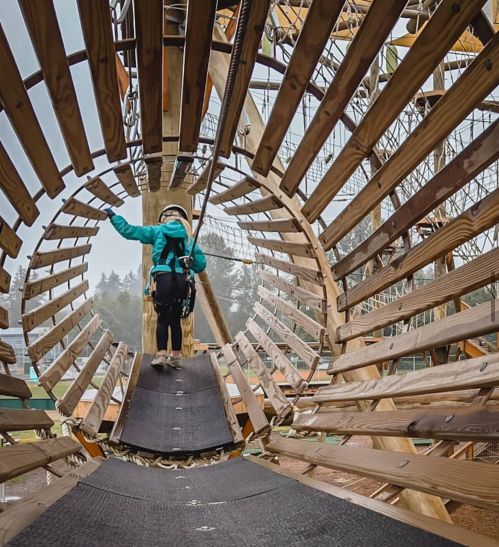A child in a harness walks through a circular wooden structure at an adventure park, surrounded by trees.