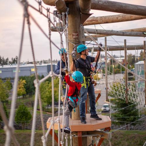 Two people in harnesses and helmets navigate a ropes course, surrounded by trees and a clear sky.