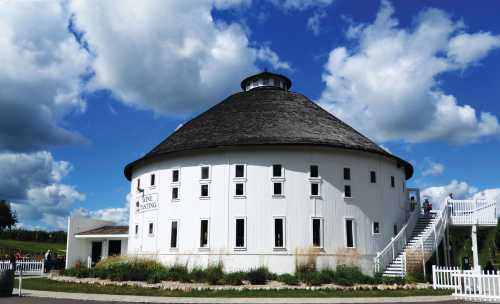 A round, white building with a circular roof, surrounded by greenery and under a blue sky with fluffy clouds.