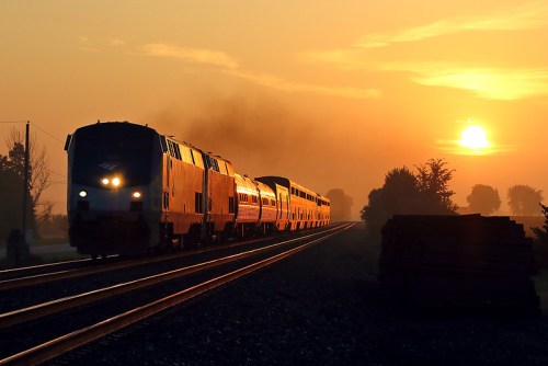 A train travels along tracks at sunset, with a warm orange glow and silhouettes of trees in the background.