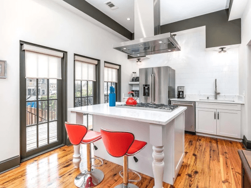 Modern kitchen with a white island, red bar stools, stainless steel appliances, and large windows for natural light.
