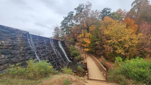A stone dam with water cascading down, surrounded by colorful autumn foliage and a wooden walkway.