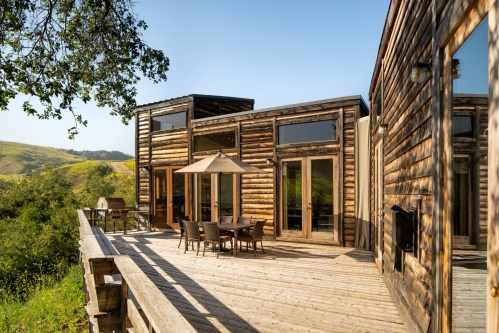 Modern wooden house with large windows and a deck, surrounded by green hills under a clear blue sky.