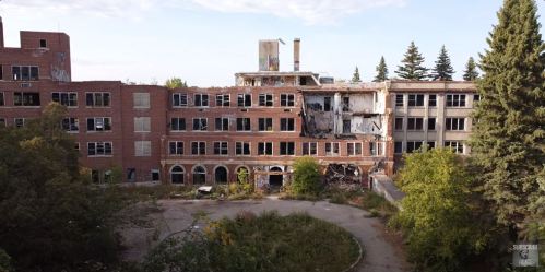 Abandoned brick building with broken windows, overgrown vegetation, and a crumbling facade under a clear sky.