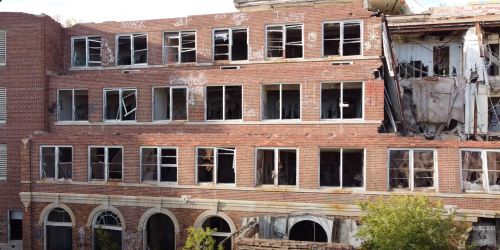 Abandoned brick building with broken windows and visible decay, surrounded by overgrown vegetation.