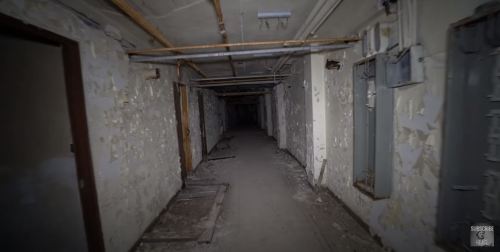 Dimly lit, abandoned hallway with peeling walls and debris on the floor, creating an eerie atmosphere.