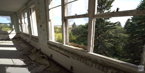 Abandoned room with large windows, peeling walls, and a view of trees outside. Sunlight filters through the dust.