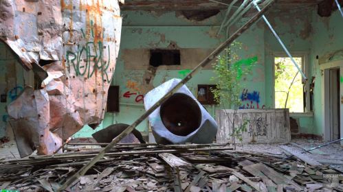 An abandoned room with peeling green walls, debris, and a large concrete sculpture amidst overgrown plants.
