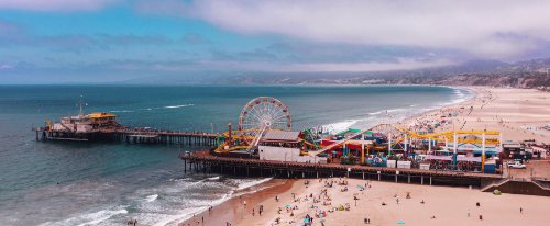 Aerial view of a vibrant pier with a ferris wheel, beachgoers, and a sandy shoreline under a cloudy sky.