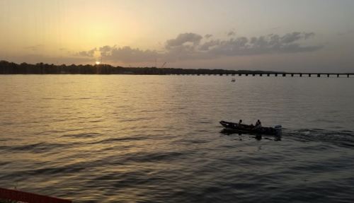 A serene sunset over a calm lake, with a small boat gliding across the water and a distant bridge in the background.