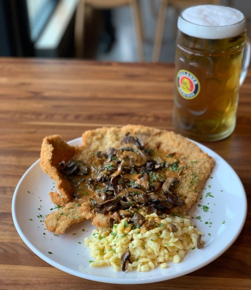 A plate of breaded schnitzel topped with mushrooms, served with spaetzle and a glass of beer on a wooden table.