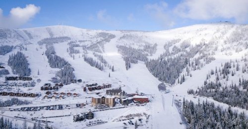 Aerial view of a snowy mountain resort with ski slopes and lodges surrounded by evergreen trees.