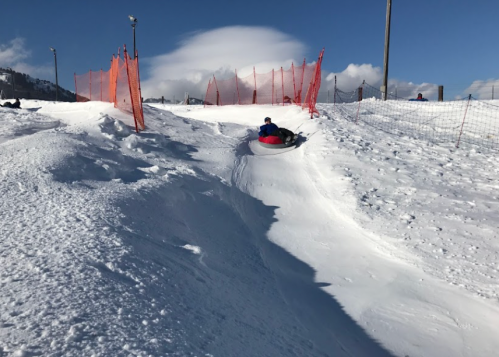 A person sledding down a snowy hill, surrounded by orange safety fencing and a clear blue sky.