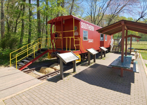 A red train caboose labeled "Southern X441" beside a picnic area with benches and informational signs in a green park.
