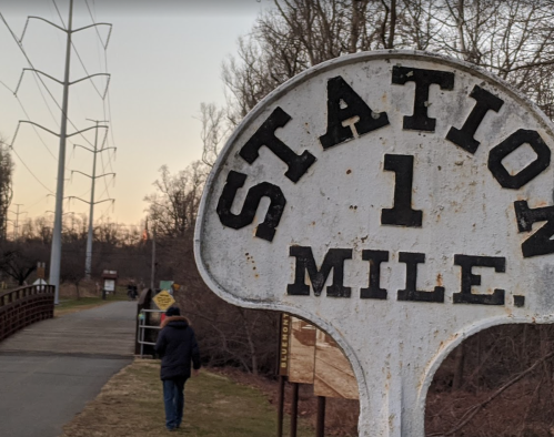 A weathered sign reading "Station 1 Mile" near a walking path, with a person walking in the background.