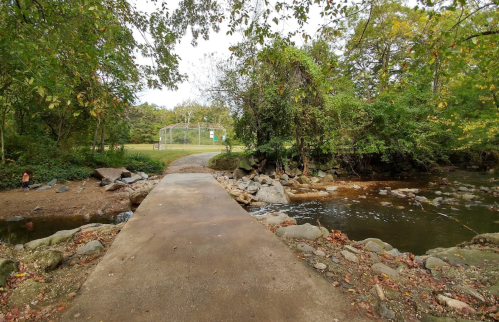 A serene pathway leads over a small stream, surrounded by lush greenery and trees, with a grassy area in the background.