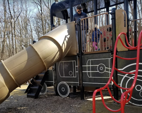 A playground with a large slide and children playing, surrounded by trees in a park setting.