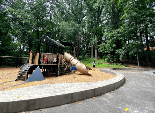 A playground featuring a large slide shaped like a train, surrounded by trees and gravel. Children are playing nearby.
