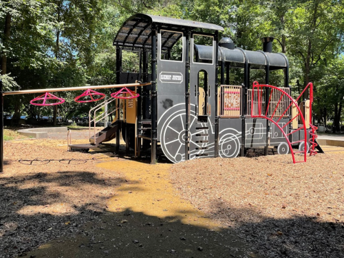 A playground structure designed like a black train, featuring slides and climbing areas, surrounded by trees.