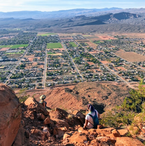 Two hikers sit on a rocky ledge overlooking a valley and town below, with mountains in the distance under a clear sky.