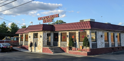 Exterior of Metuchen Diner with a red roof, stone walls, and a parking lot under a blue sky with scattered clouds.