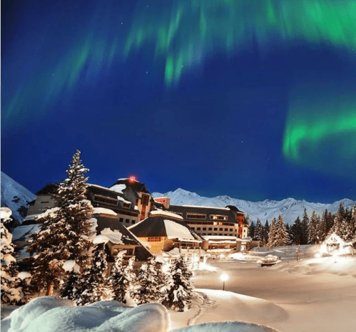 A snowy landscape with a hotel, surrounded by trees, under a vibrant aurora borealis in a night sky.