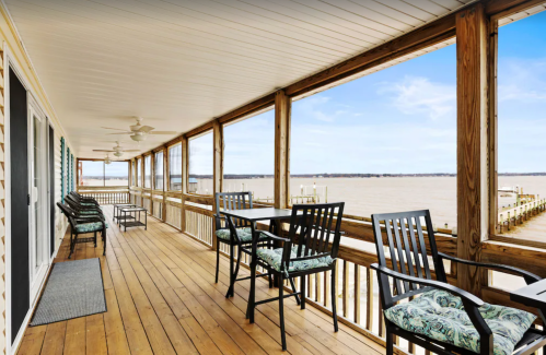 A wooden balcony with chairs and tables overlooking a calm body of water and a clear blue sky.