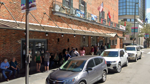 A busy street scene with a brick building, people waiting outside, and parked cars along the curb.