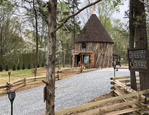 A whimsical hobbit house with a round wooden structure, surrounded by trees and a gravel path, marked by a sign.