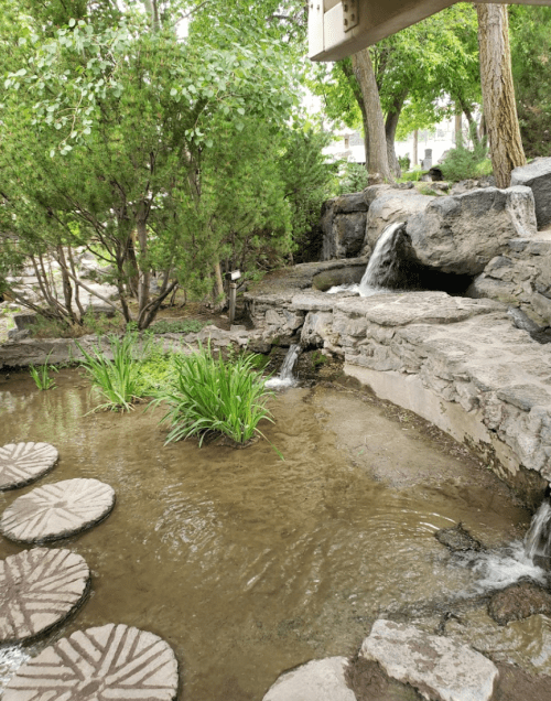 A serene garden scene featuring a small pond, stepping stones, and cascading waterfalls surrounded by greenery.