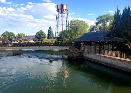 A serene view of a river with a wooden bridge, a pagoda-style structure, and a water tower in the background.