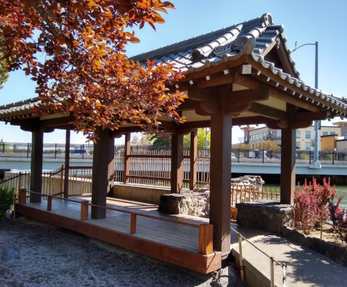 A wooden pavilion with a tiled roof surrounded by colorful foliage and a calm waterway in the background.