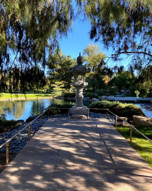 A stone lantern stands on a pathway by a serene pond, surrounded by lush greenery and blue skies.
