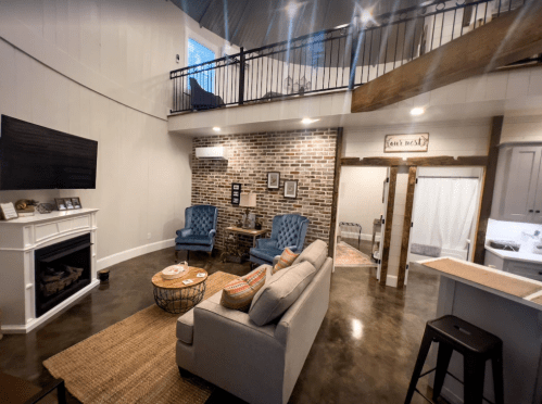 Cozy living room with a sofa, two blue chairs, a brick wall, and a staircase leading to a loft area.