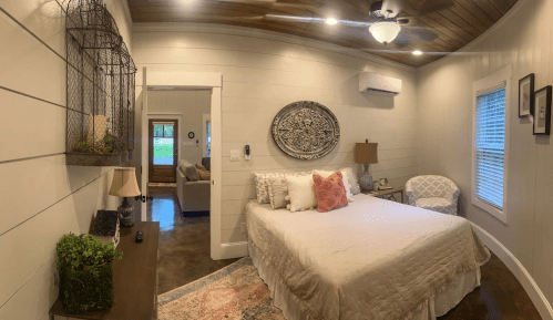 Cozy bedroom with a bed, decorative wall art, a chair, and natural light from windows. Warm, inviting atmosphere.